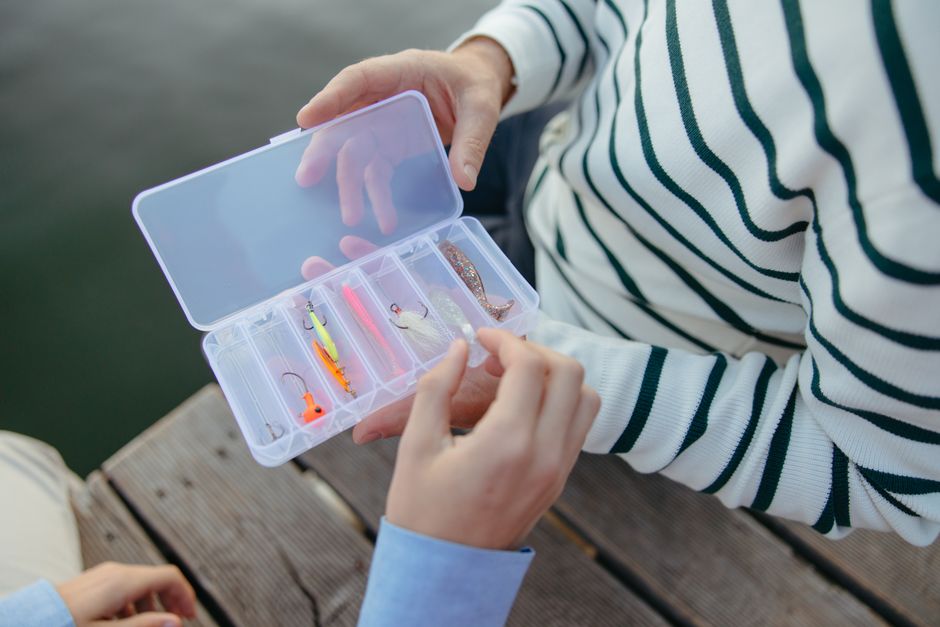 Hands selecting colorful fishing lures from a tackle box on a wooden dock.