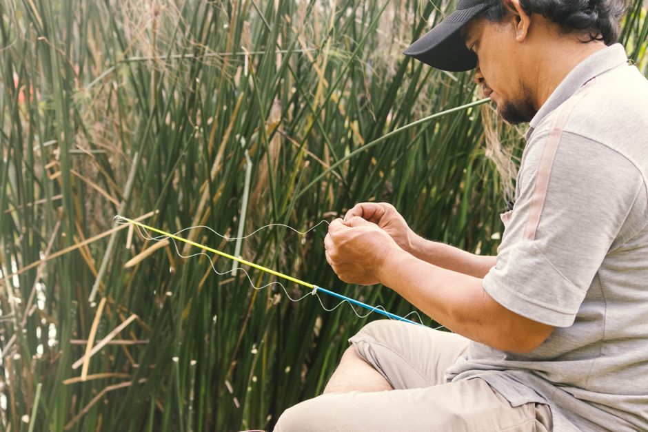 Man carefully ties a fishing line while sitting by tall grasses, enjoying a peaceful moment outdoors.